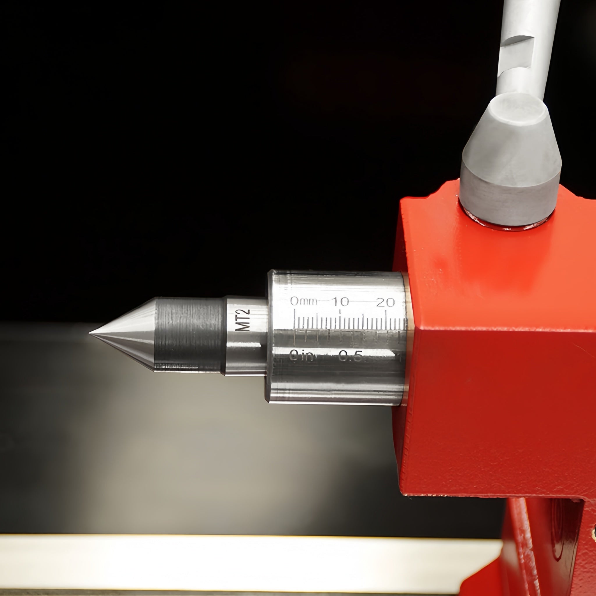 Close-up of a precision machined metal dial indicator or measuring tool with a pointed silver tip, a silver scale barrel marked 0-10-20 mm, and a bright red mounting block against a dark background.