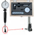 A dial indicator set shown with the indicator gauge detached on the left, the gauge mounted inside a wooden storage case on the upper right, and the mounting clamp on the lower right with a red arrow pointing to the feet. The product is photographed on a plain white background.