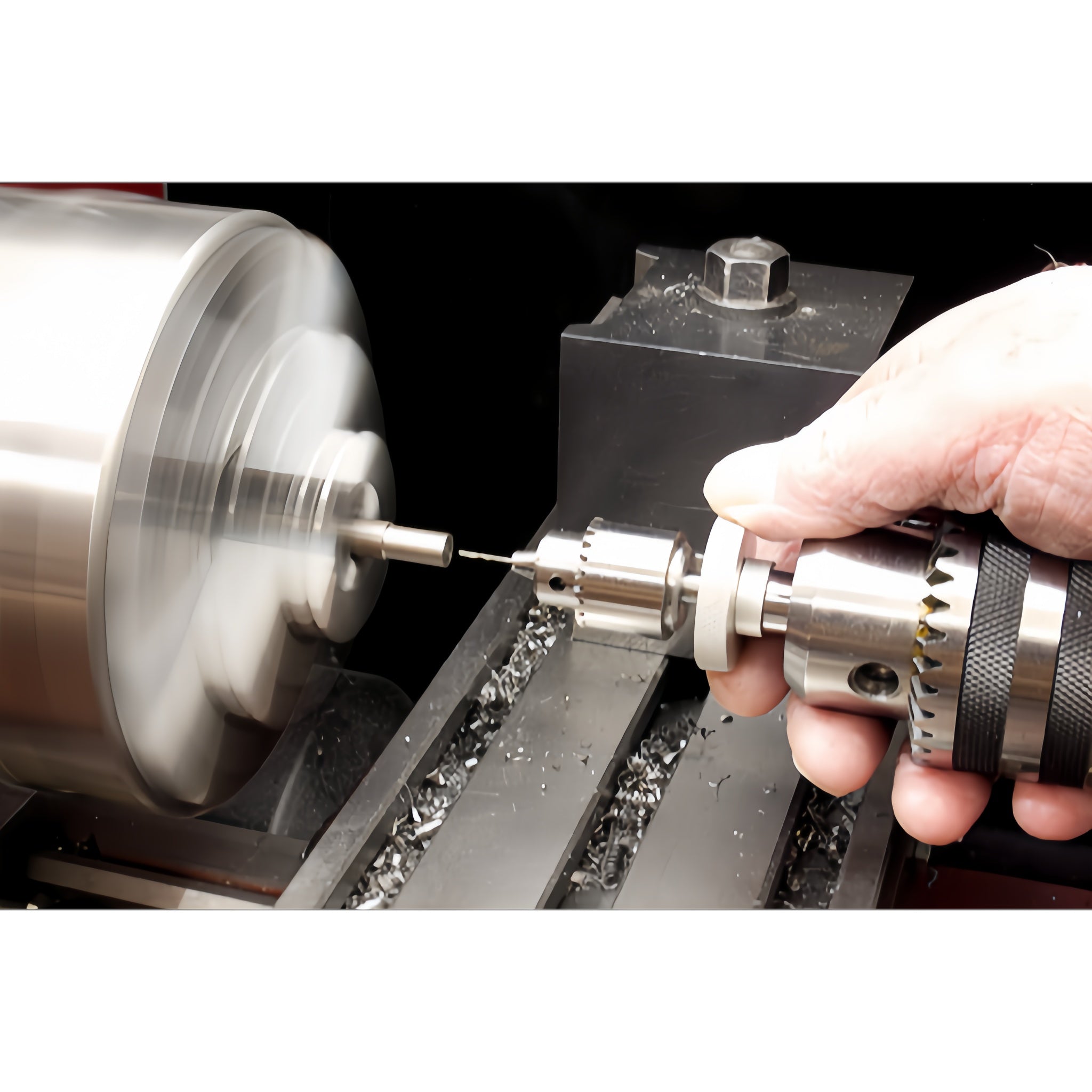 A close-up of a metal lathe machine in operation, with a spinning silver workpiece on the left and a hand holding a drill chuck on the right, showing metal shavings being produced.