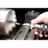 A close-up of a metal lathe machine in operation, with a spinning silver workpiece on the left and a hand holding a drill chuck on the right, showing metal shavings being produced.