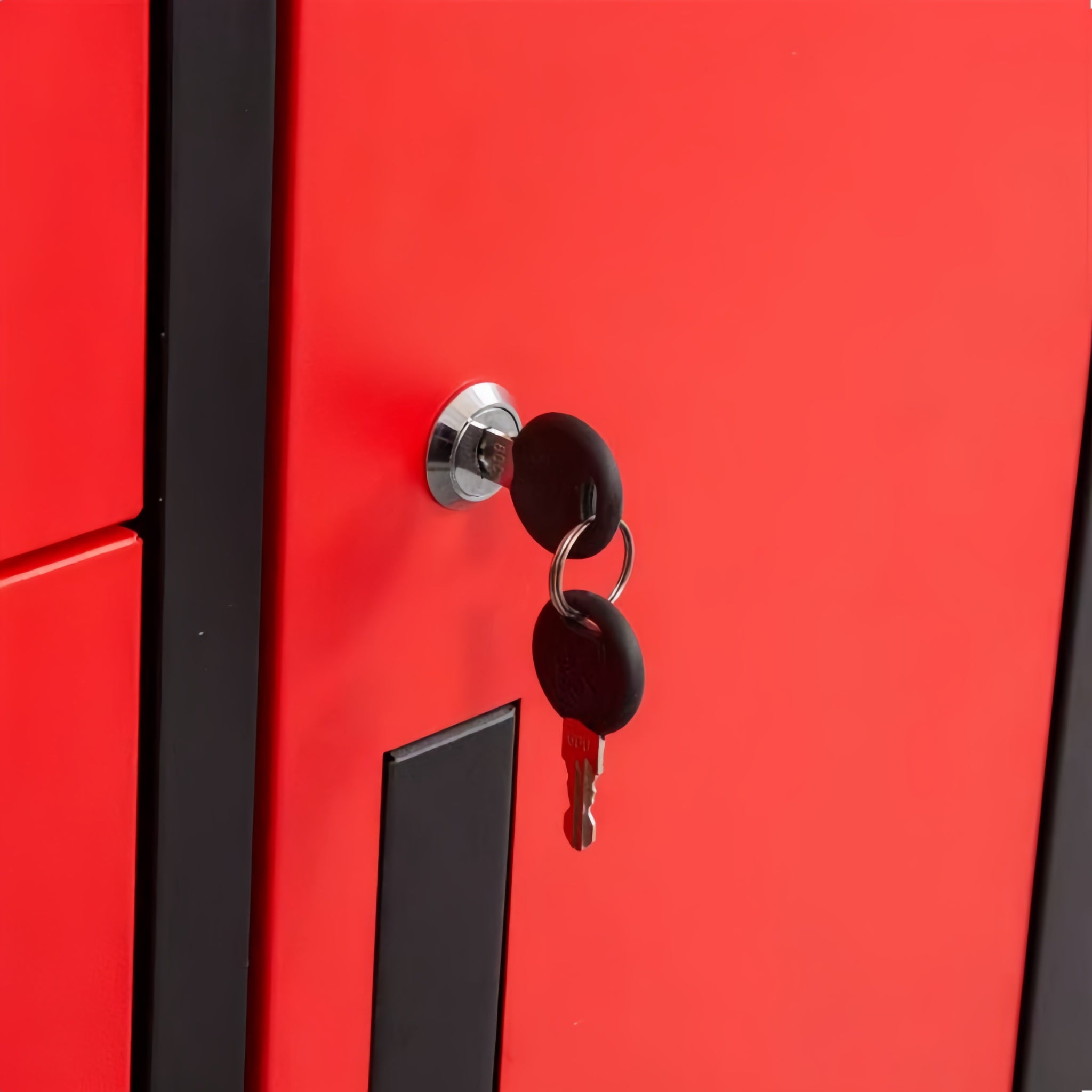 Close-up of a bright red metal locker or cabinet door with a black vertical trim strip, a silver lock cylinder, two black keys inserted, and a black rectangular handle plate.