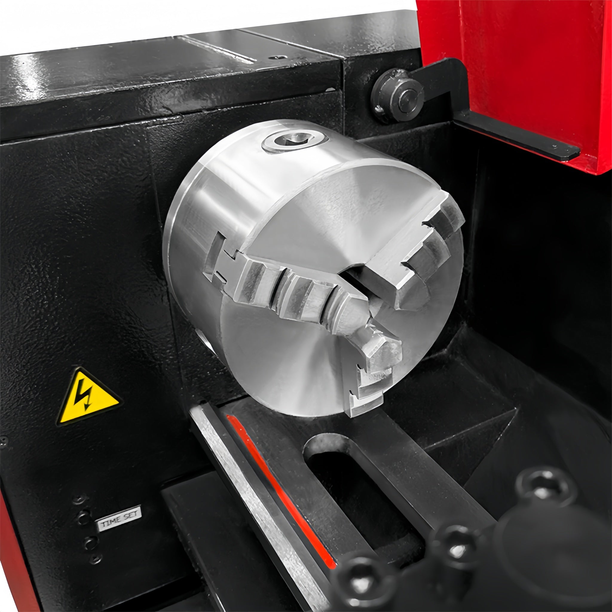 A close-up of a metallic three-jaw lathe chuck mounted on a black and red industrial machine, with a yellow warning label visible on the side.