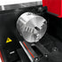 A close-up of a metallic three-jaw lathe chuck mounted on a black and red industrial machine, with a yellow warning label visible on the side.