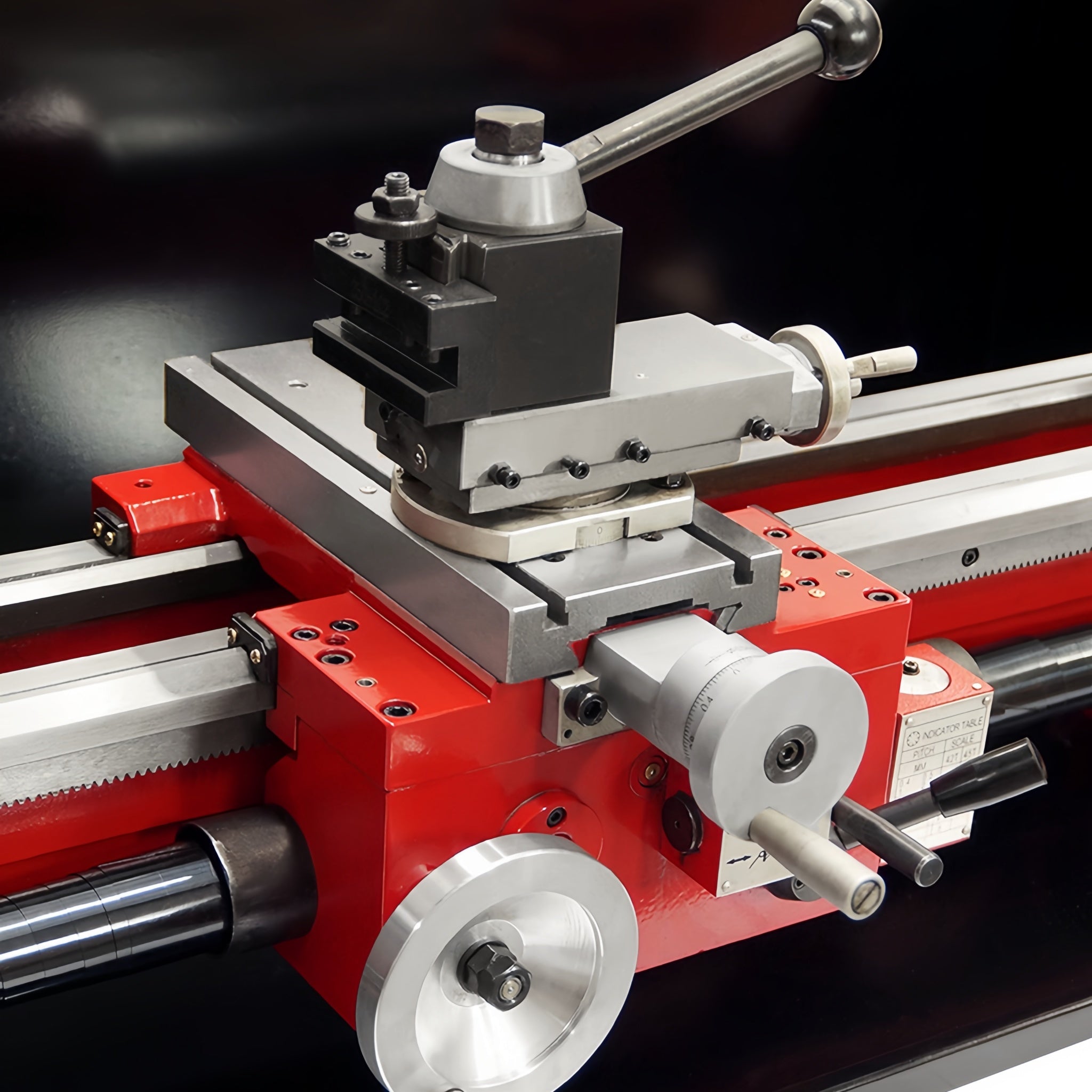 Close-up of a red and silver metal lathe machine component with a carriage, quick change tool post, and a handwheel on a dark background.