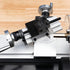 Close-up of a precision optical or laser alignment device with silver and black metal components, a threaded adjustment screw, a white measuring scale card, and a light wood background.