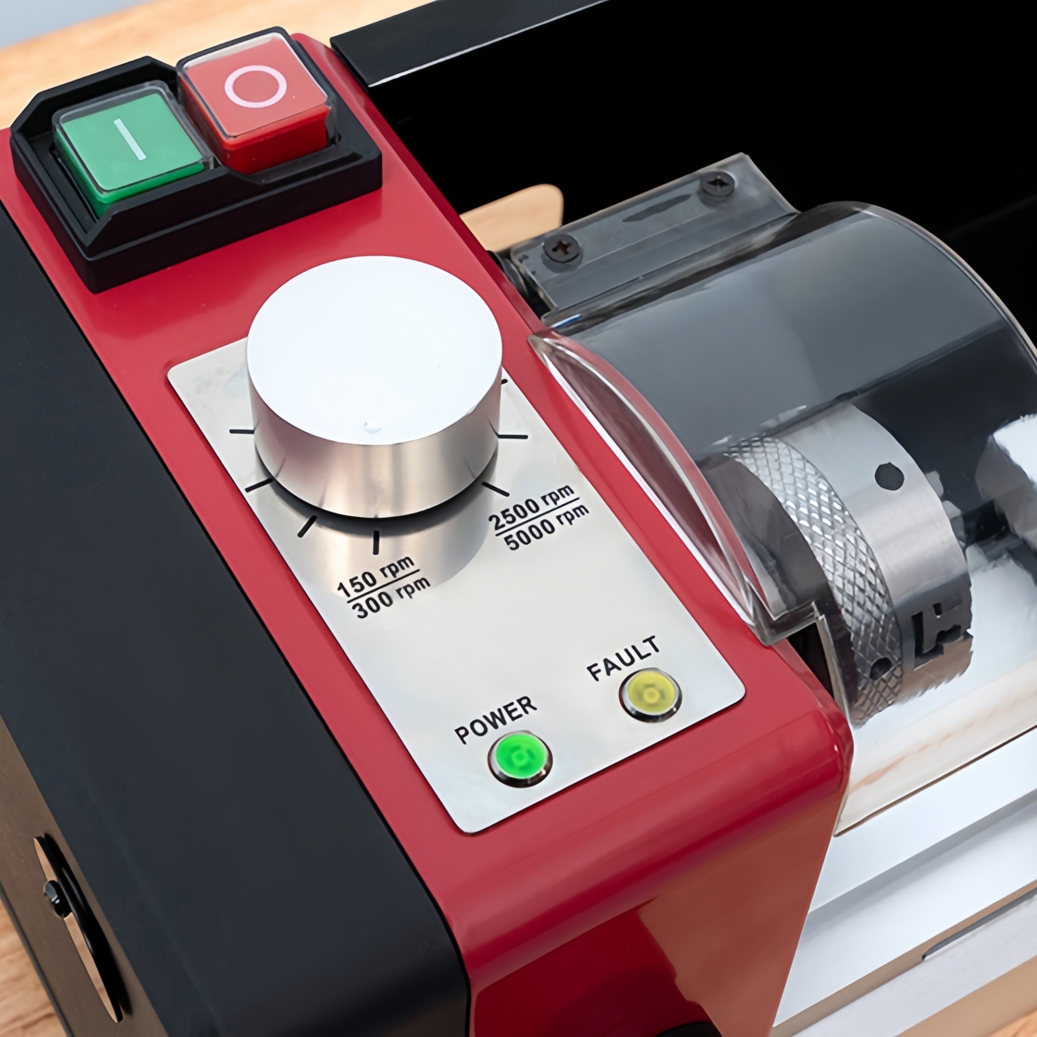 Close-up of a red and black industrial machine control panel with a large silver speed dial, green and red buttons, and a transparent protective cover over a metal roller mechanism.