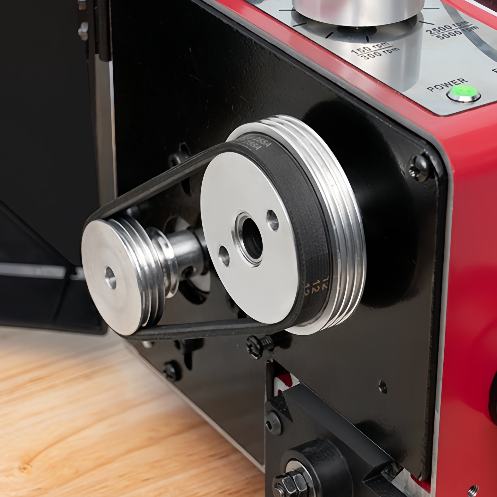 Close-up of a red and black industrial pulley system with metallic belts and pulleys, mounted on a machine frame with a wood surface below.