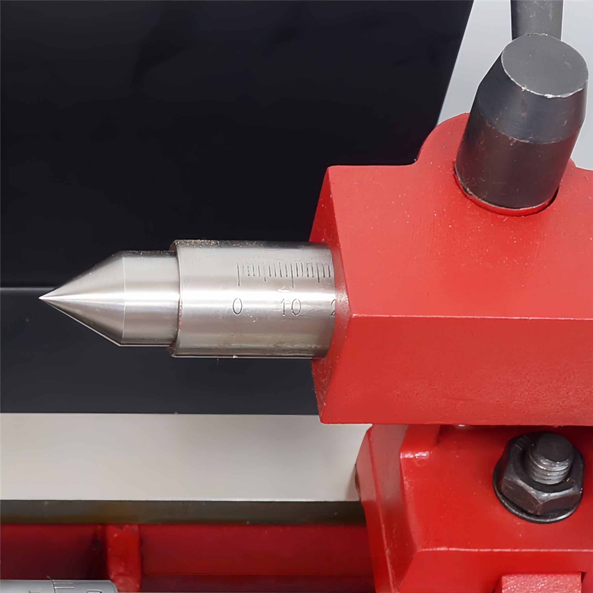 Close-up of a machined metal dial indicator or precision measuring tool with a pointed silver tip and engraved scale, mounted on a bright red clamp or fixture.