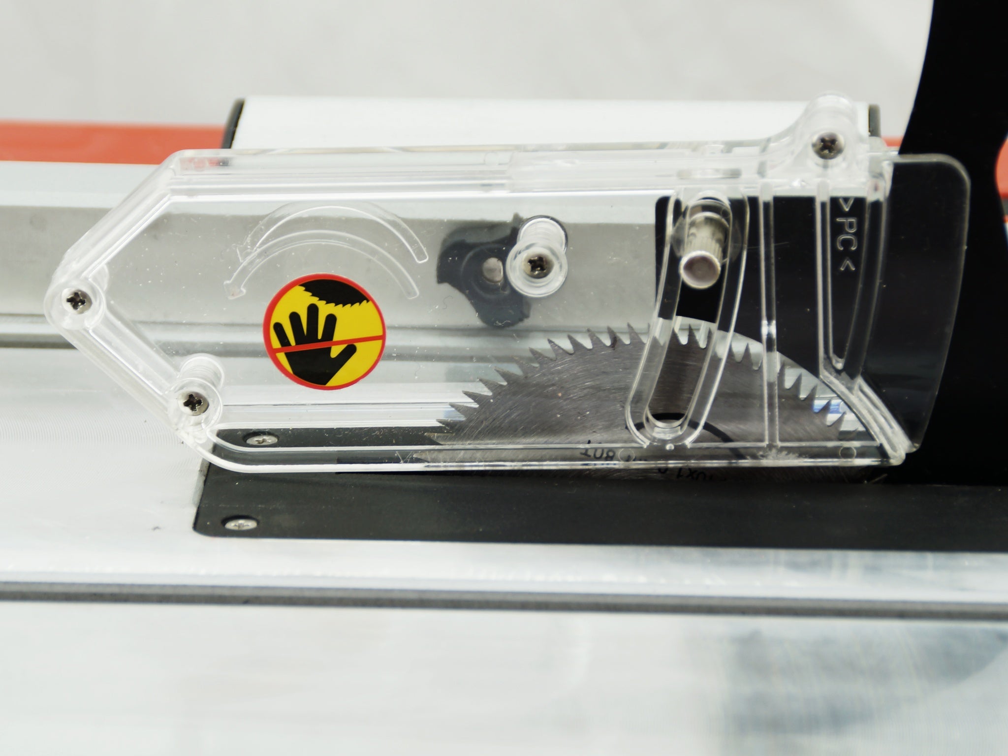 Close-up of a table saw blade guard with a clear plastic cover, black metal components, and a yellow and red warning sticker showing a hand inside a circle.