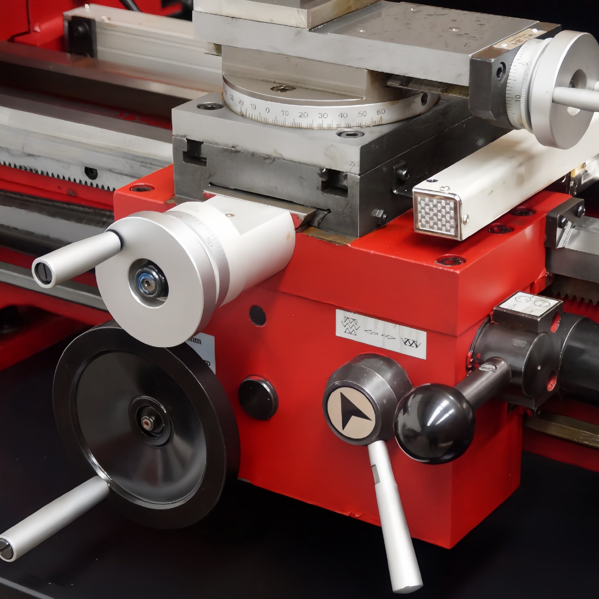 Close-up of a red and silver machine component with handwheels, knobs, and metal adjustment parts on a dark surface.