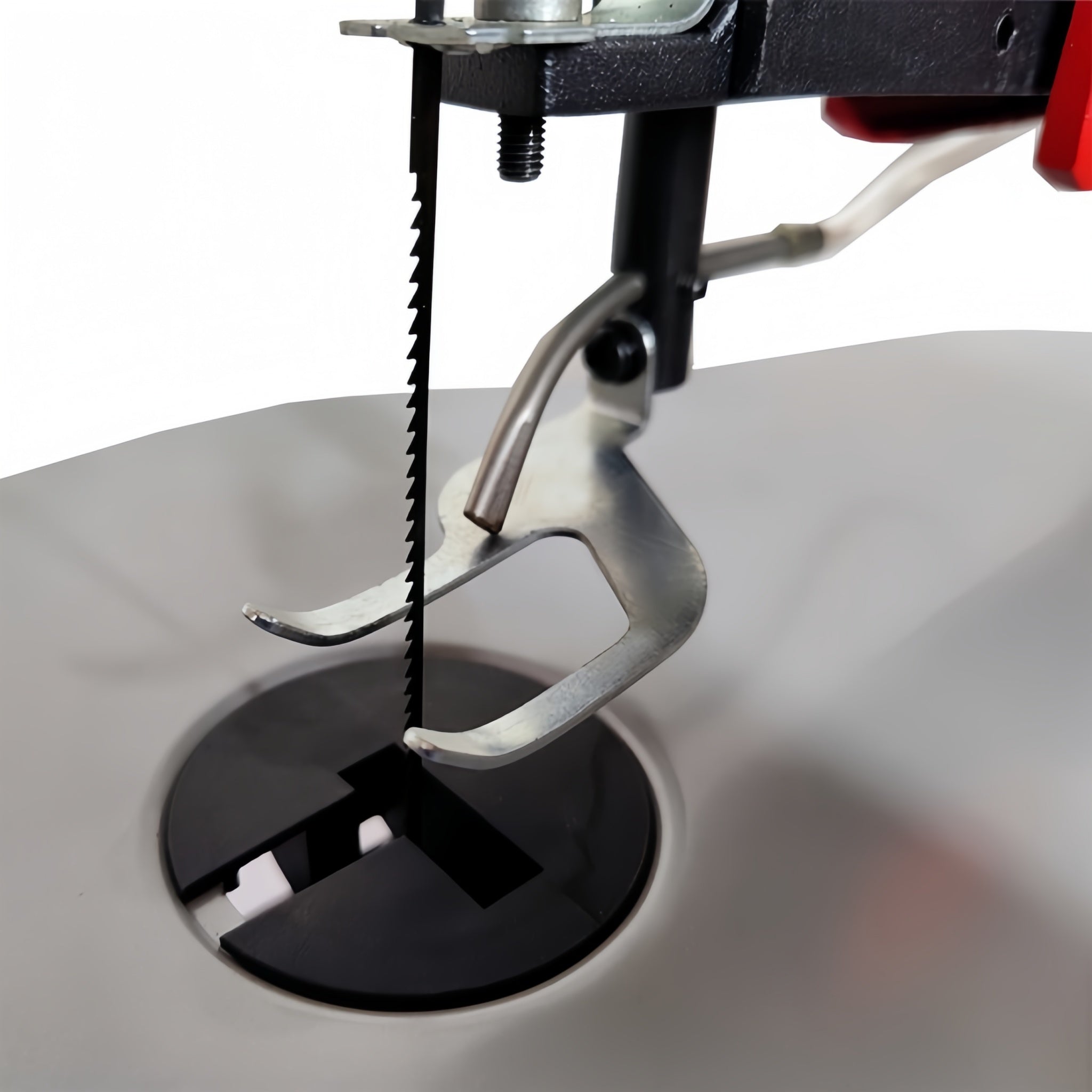 Close-up of a scroll saw blade and presser foot assembly over a circular black opening, with a white fabric surface below on a clean white background.