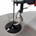 Close-up of a scroll saw blade and presser foot assembly over a circular black opening, with a white fabric surface below on a clean white background.